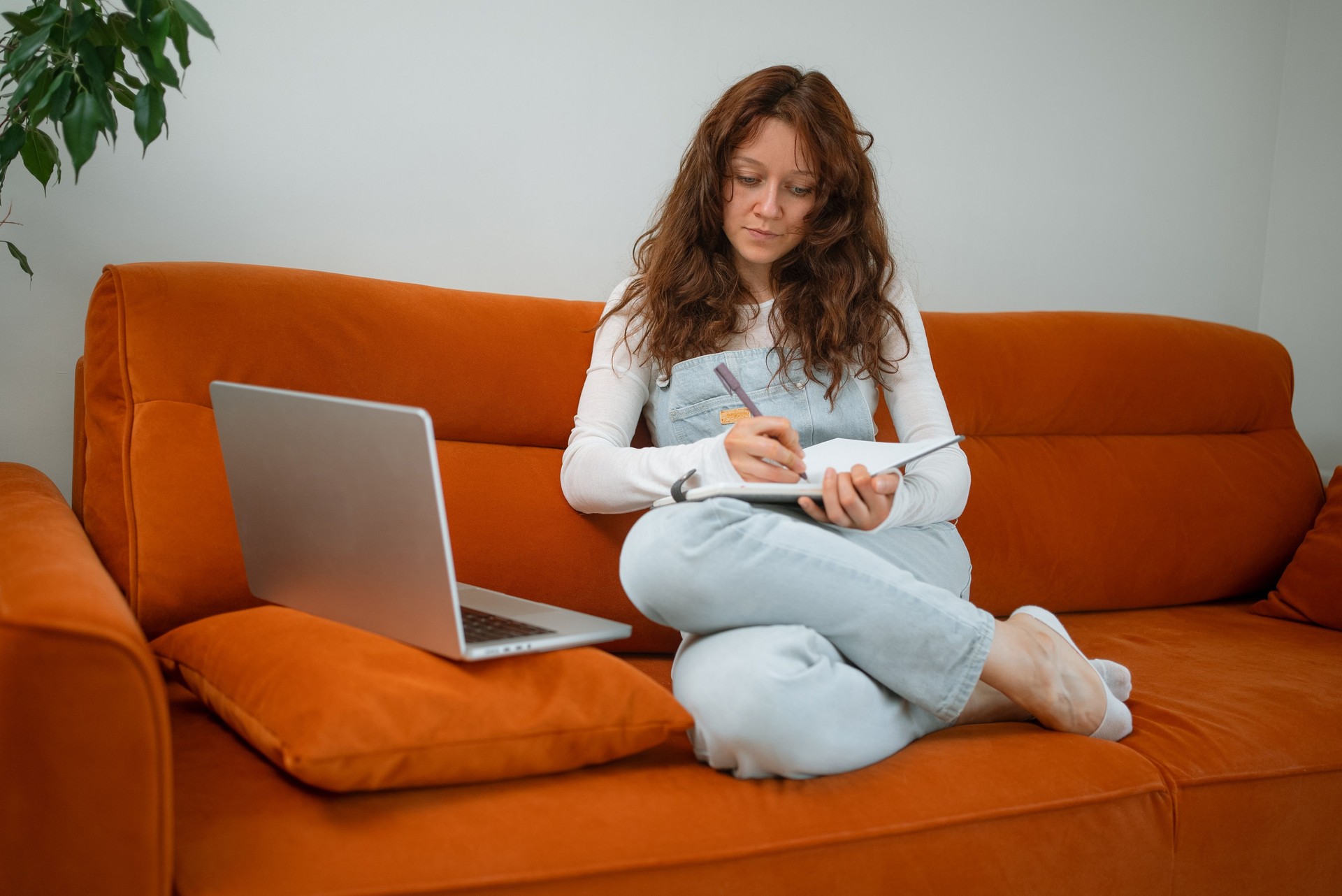 Remote freelancer writing notes in notebook, working on laptop from cozy orange sofa, enjoying flexible schedule and comfortable home workspace